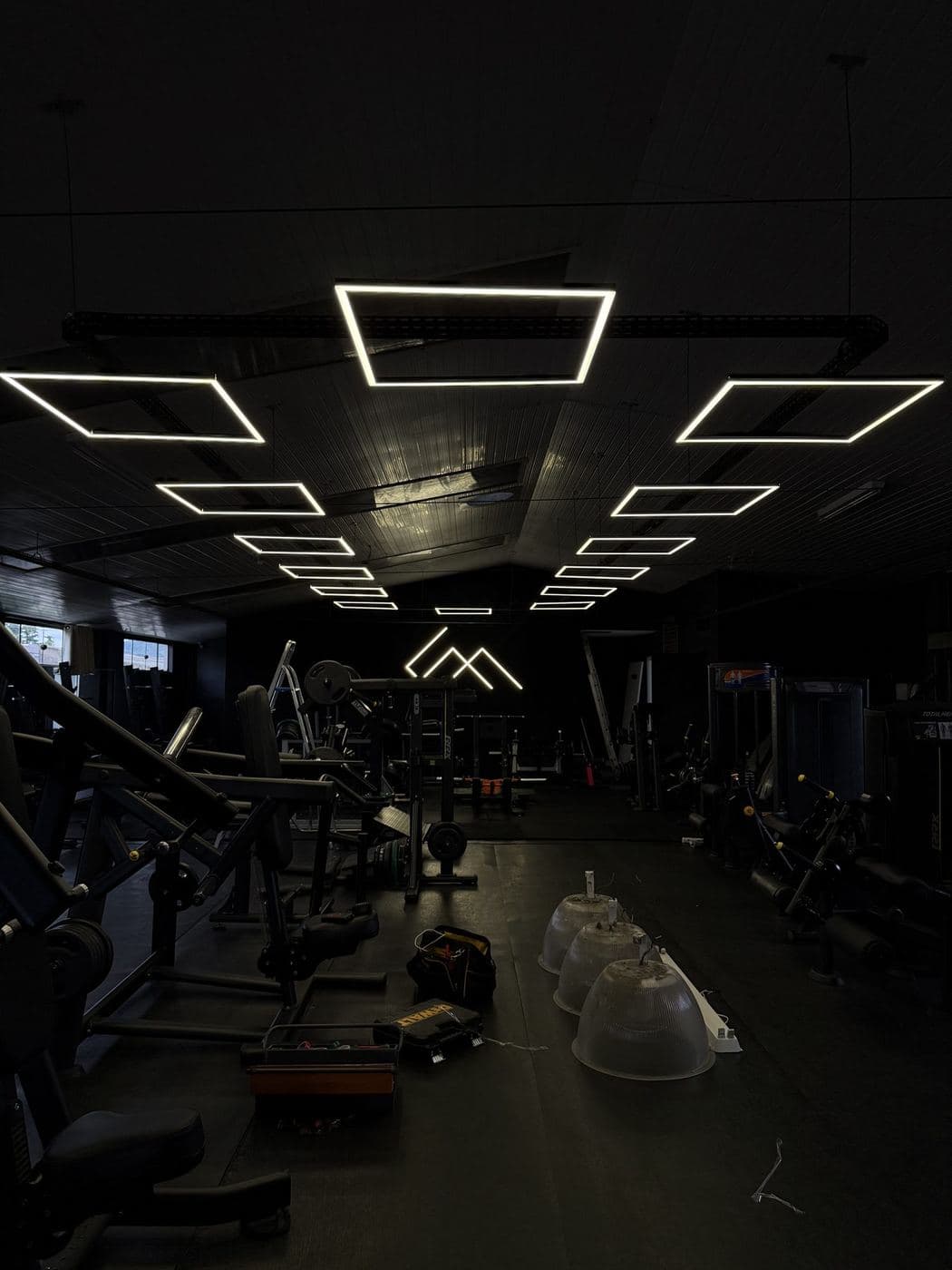 Academia em montagem: Luminárias de perfil de LED em formato de quadros sendo montadas no teto da área de treino.