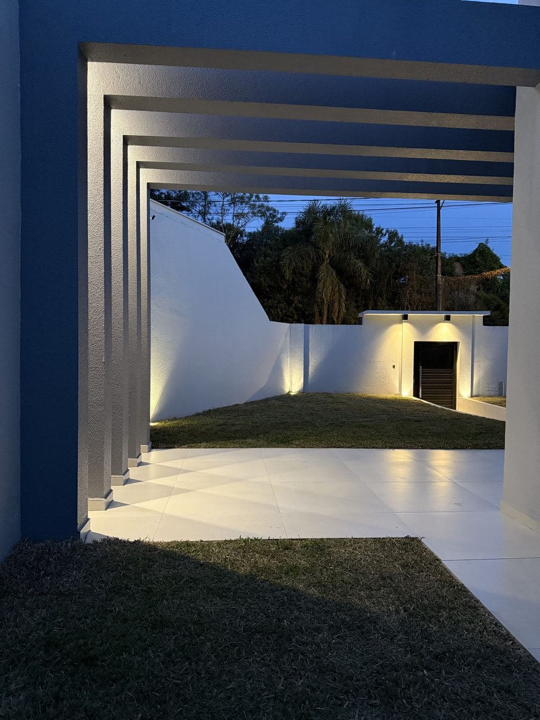 Área de lazer: Luz rasante nos muros, arandelas e iluminação sob o pergolado.