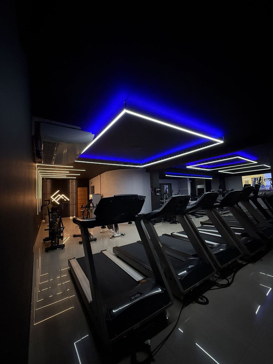 Academia, área de esteiras: Sanca com LED azul, quadros de perfil de LED no forro e linhas de luz no piso.
