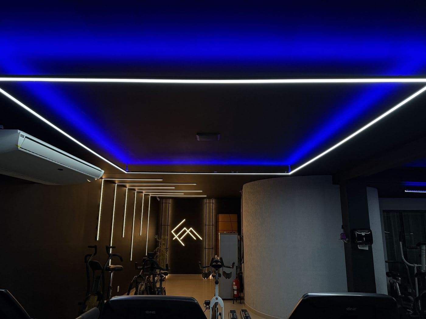 Academia, área de cardio: Sanca azul com perfil de LED branco no forro e linhas verticais na parede do fundo.