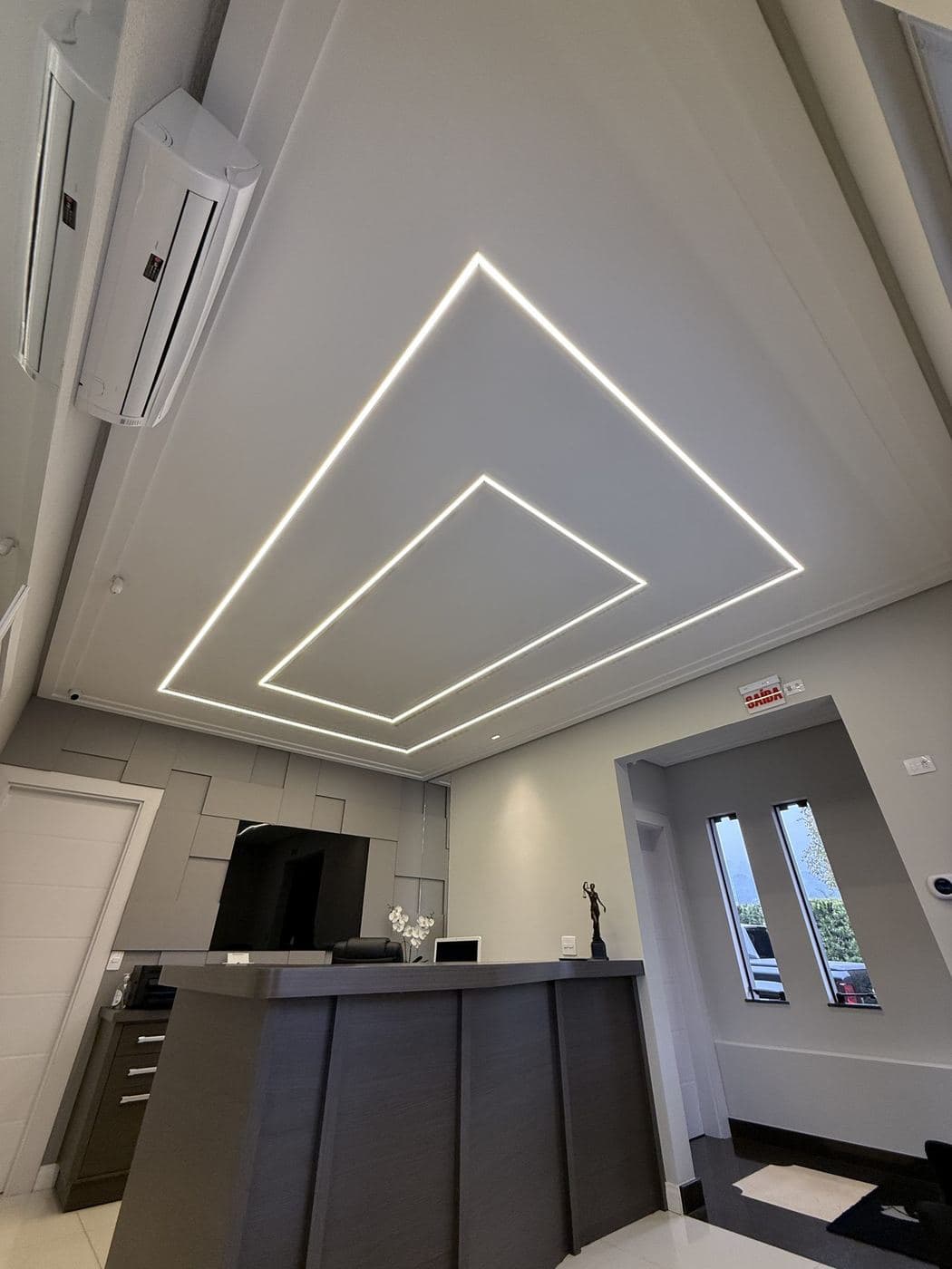 Recepção: Perfis de LED em quadrados concêntricos no forro sobre o balcão de atendimento.